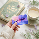 Hand presenting nebula violin card skin beside The Enchanted Symphony book and tea BY THE STICKER ERA