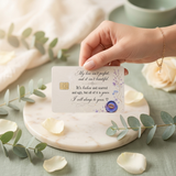 A hand holdsing a printed  credit card skin card with a chip, cursive text, and a blue wax seal, styled on a marble coaster with eucalyptus leaves and rose petals.