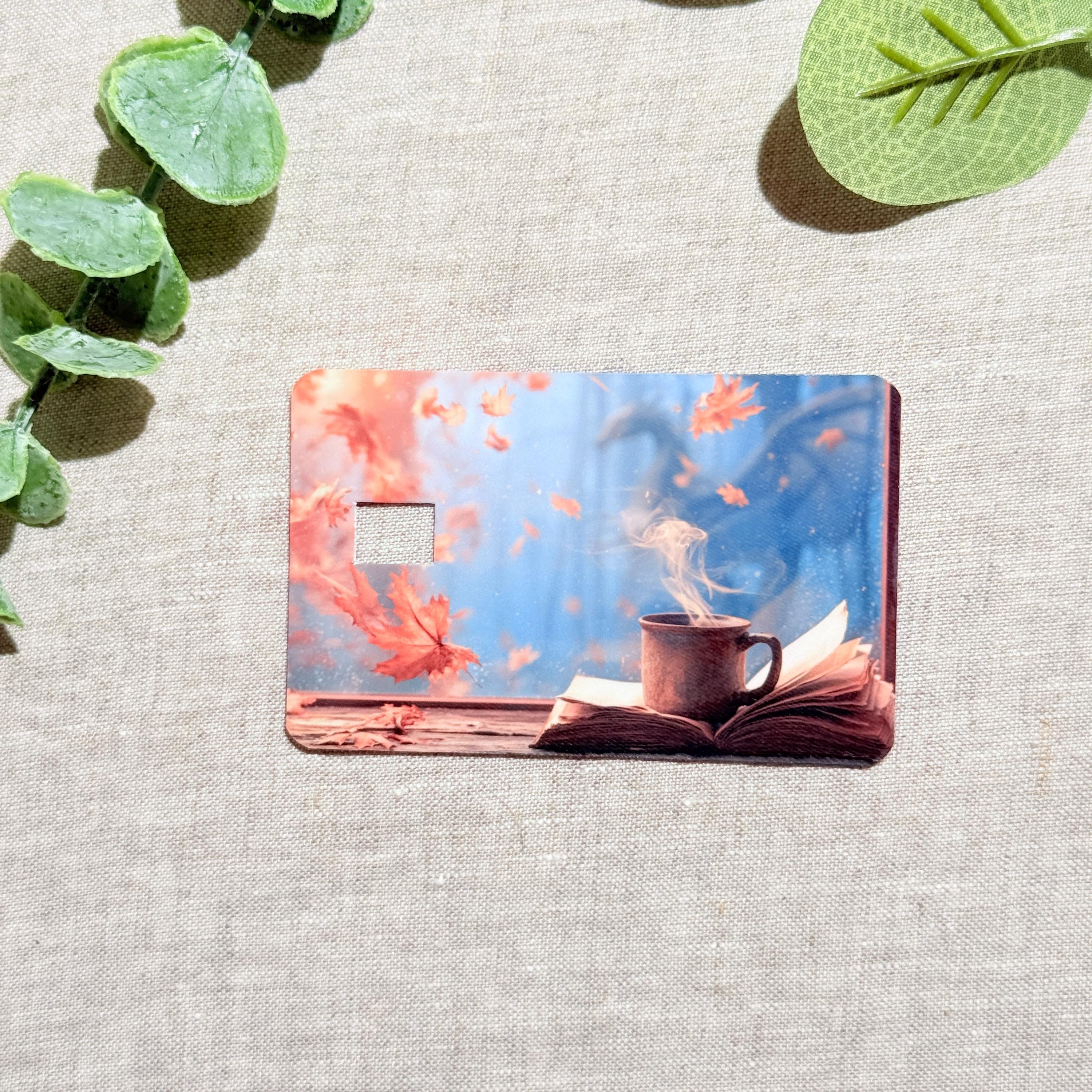Fall card skin with falling leaves, a steaming coffee mug on an open book and a dragon in the blue mist on a debit card by The Sticker Era