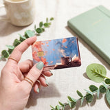 Hand holding the fall cozy books and coffee card skin beside a sage notebook by The Sticker Era