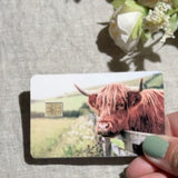 Hand holding the highland cow meadow debit card skin over linen and white flowers by The Sticker Era