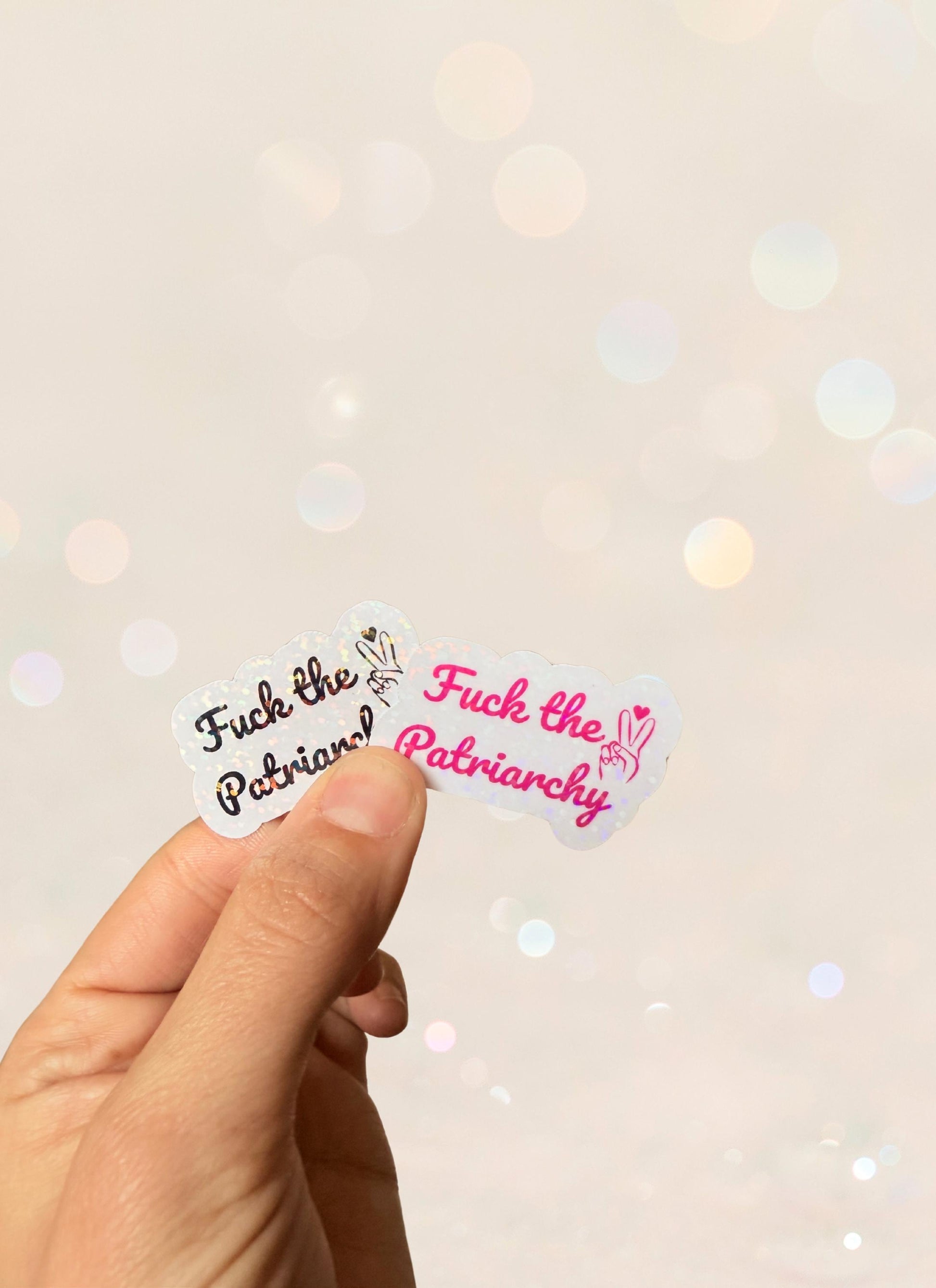 Hand holding a small sticker that reads “F*ck the Patriarchy” in black and pink script. Ideal for feminists, bold statement pieces, and empowerment gifts.