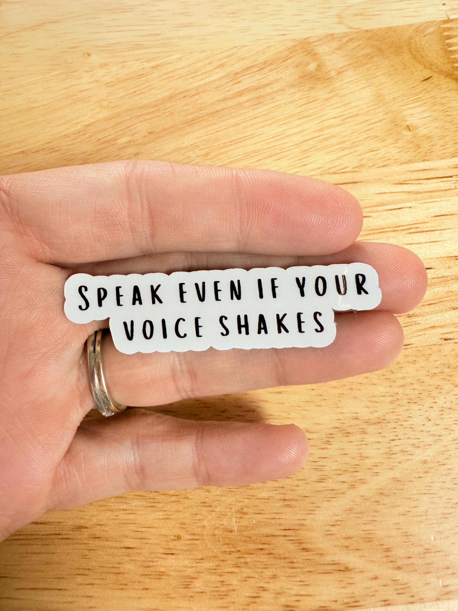 Close-up of the "Speak Even If Your Voice Shakes" sticker held near a person's lips