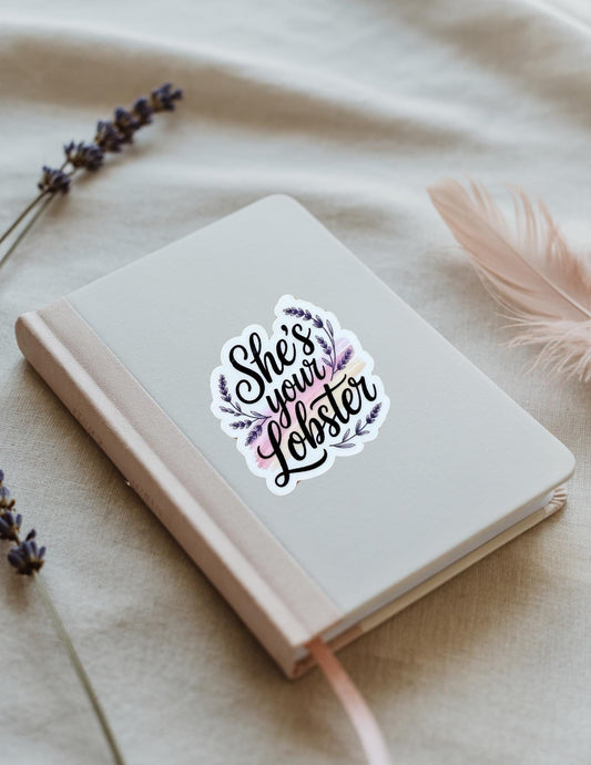 Vinyl sticker with the phrase “She’s Your Lobster” in black script surrounded by purple leaves, shown on a beige notebook. Inspired by the TV show Friends and perfect for romantic gifts or fans.