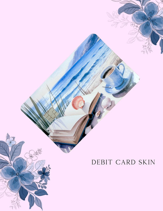 A debit card skin featuring a beachy aesthetic design is displayed on a pink background with floral accents. The card shows an open book, a cup of coffee, seashells, and ocean waves, evoking a calm, coastal vibe. The text “DEBIT CARD SKIN” appears below the design, and the overall style is soft and serene.