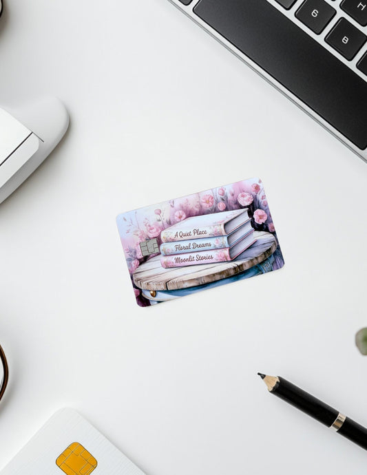 A pastel floral-themed debit card skin lies on a white desk beside a laptop, pen, and smart card reader. The card features a romantic book stack with titles "A Quiet Place," "Floral Dreams," and "Moonlit Stories," surrounded by soft pink flowers. The setup conveys a cozy, bookish vibe with a touch of elegance.