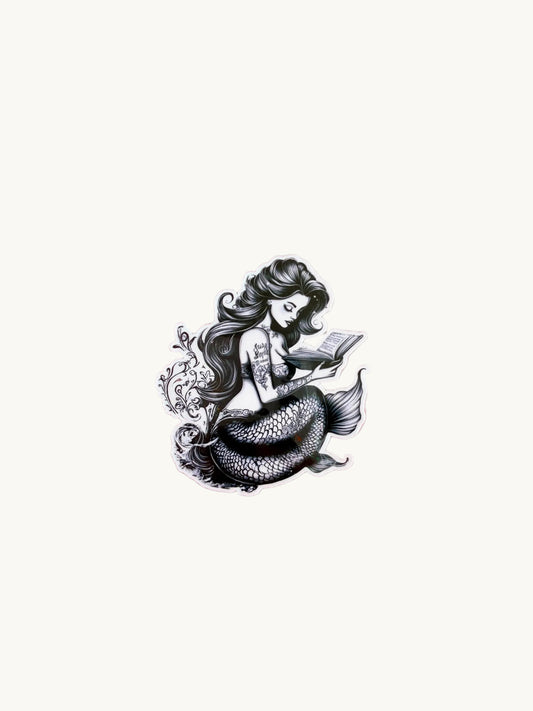 Black and white mermaid reading a book, covered in intricate tattoos. Artistic and moody fantasy-style sticker on a light background.