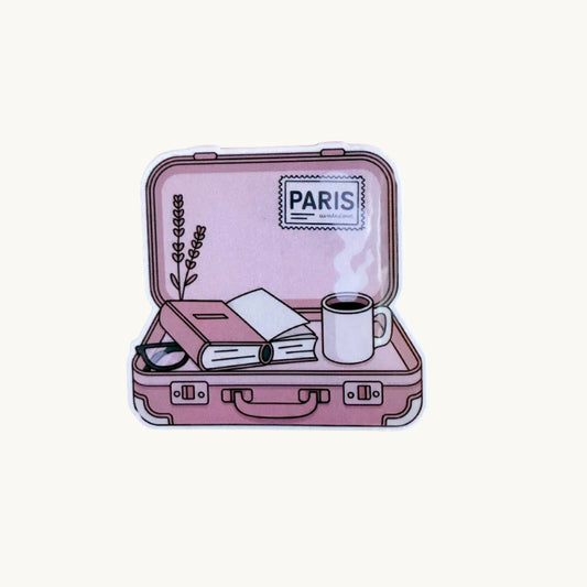 Sticker of an open pink suitcase with a book, coffee mug, sunglasses, and a 'Paris' label inside, on a soft pink background.