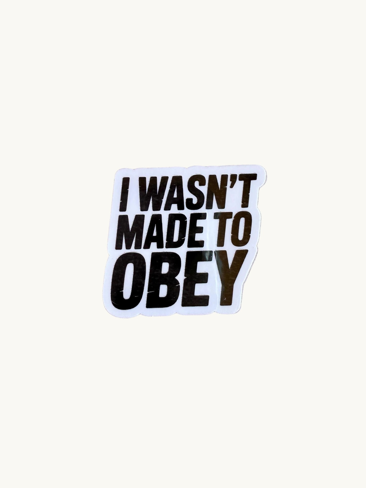 Sticker with bold black text that says 'I Wasn't Made to Obey,' placed on a round soft surface with a pink background.