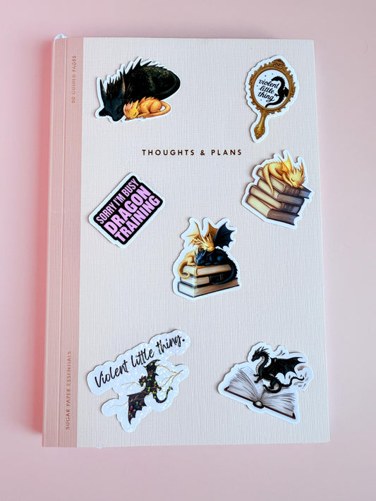 "A set of seven dragon-themed vinyl stickers displayed on a light pink background, featuring sleeping dragons, fantasy books, flying dragons, and romantasy-inspired quotes."