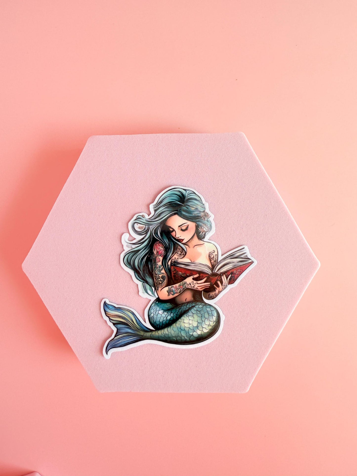 Teal-haired mermaid with green tail reading a red book. Detailed tattoos on arms. Soft fantasy-style sticker on a pink background.