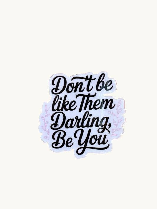 Sticker with the quote 'Don't be like them darling, be you' in black script font, with light lavender leaves around the text, held by a hand on a pink background.
