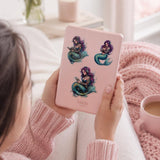 purple haired mermaid stickers, one reading, one listening to audio, and one hugging a book all on a pink kindle being held by a girl in pink with a pibk cozy blanket and coffee and pink flowers in the background

