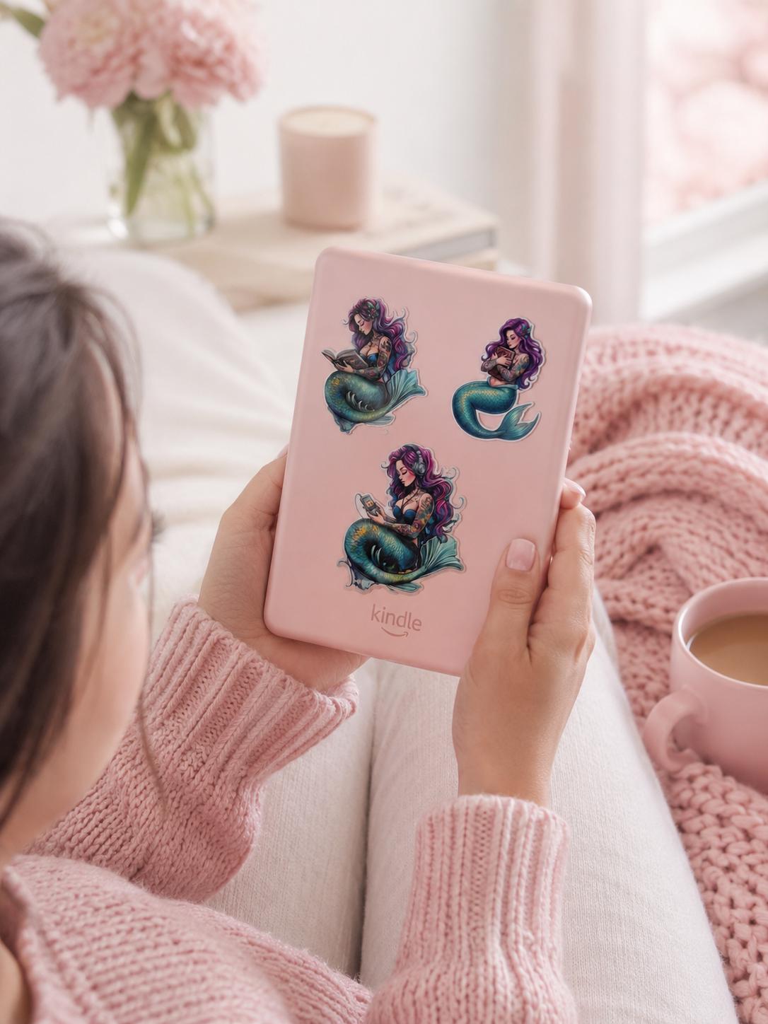 purple haired mermaid stickers, one reading, one listening to audio, and one hugging a book all on a pink kindle being held by a girl in pink with a pibk cozy blanket and coffee and pink flowers in the background
