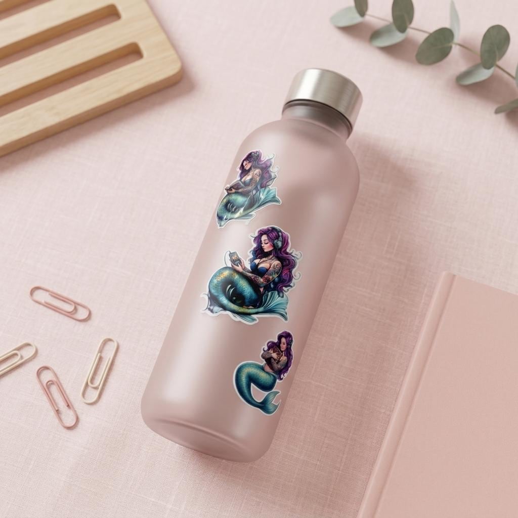 purple haired mermaid stickers, one reading, one listening to audio, and one hugging a book placed on a glass water bottle laying on a desk with pink closed journal
by the sticker era