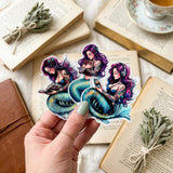 purple haired mermaid stickers, one reading, one listening to audio, and one hugging a book being held in a womans hand with sage, a brown journal and a tea cup in the background
by the sticker era