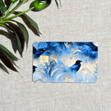 Black raven perched among blue and cream wheat botanical illustration on a waterproof debit card skin, styled flat over linen with fresh olive branches BY THE STICKER ERA
