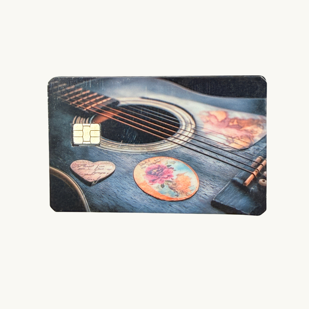 Acoustic guitar rose debit card skin with vintage blue guitar strings heart pick and painted rose soundhole on wood desk with succulent keys and guitar pick by The Sticker Era
