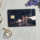 Nightcap card skin on a debit card, pocket watch and cut glass on dark marble with an open book