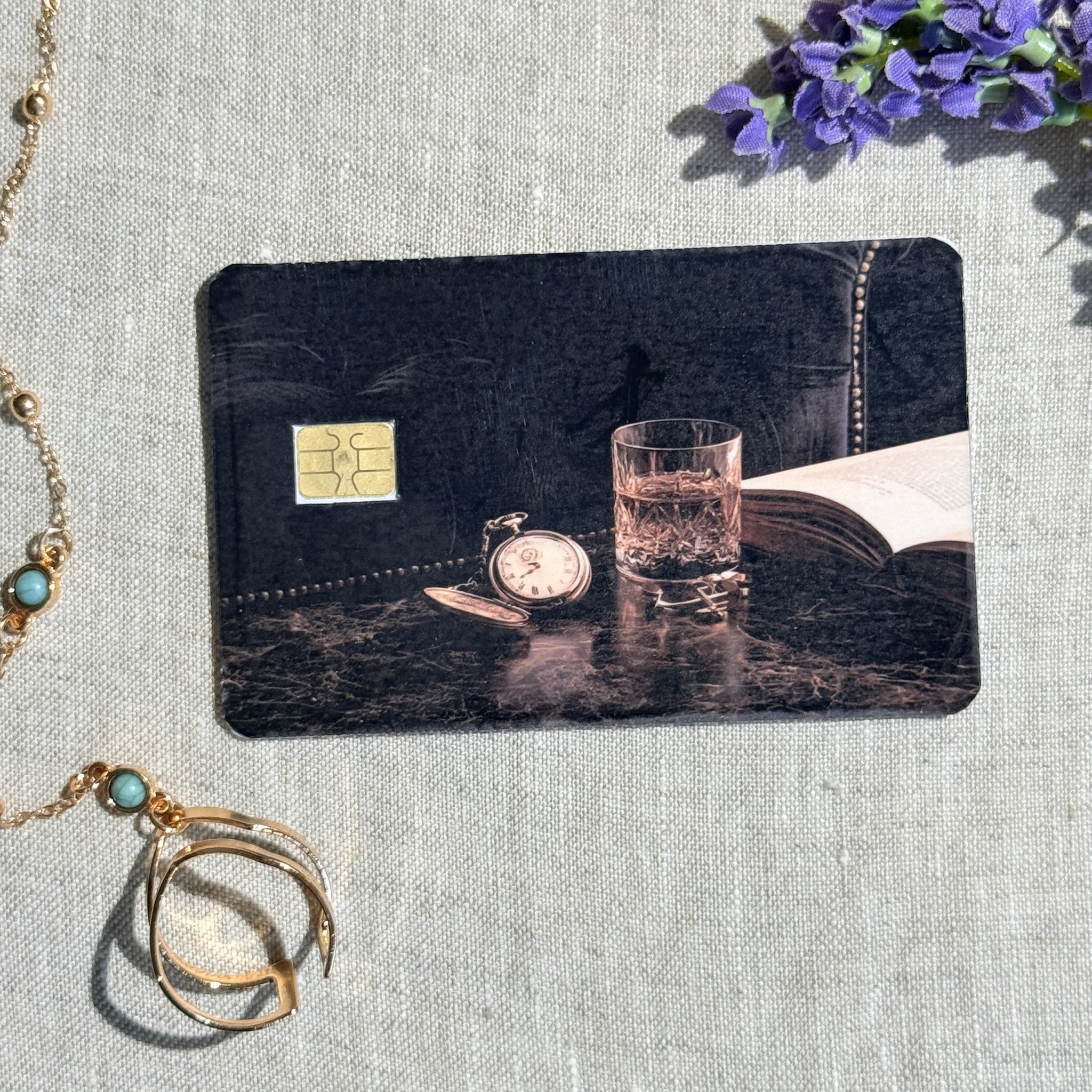 Nightcap card skin on a debit card, pocket watch and cut glass on dark marble with an open book