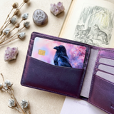 Raven cherry blossom card skin tucked into a deep purple wallet beside a vintage nature book by The Sticker Era